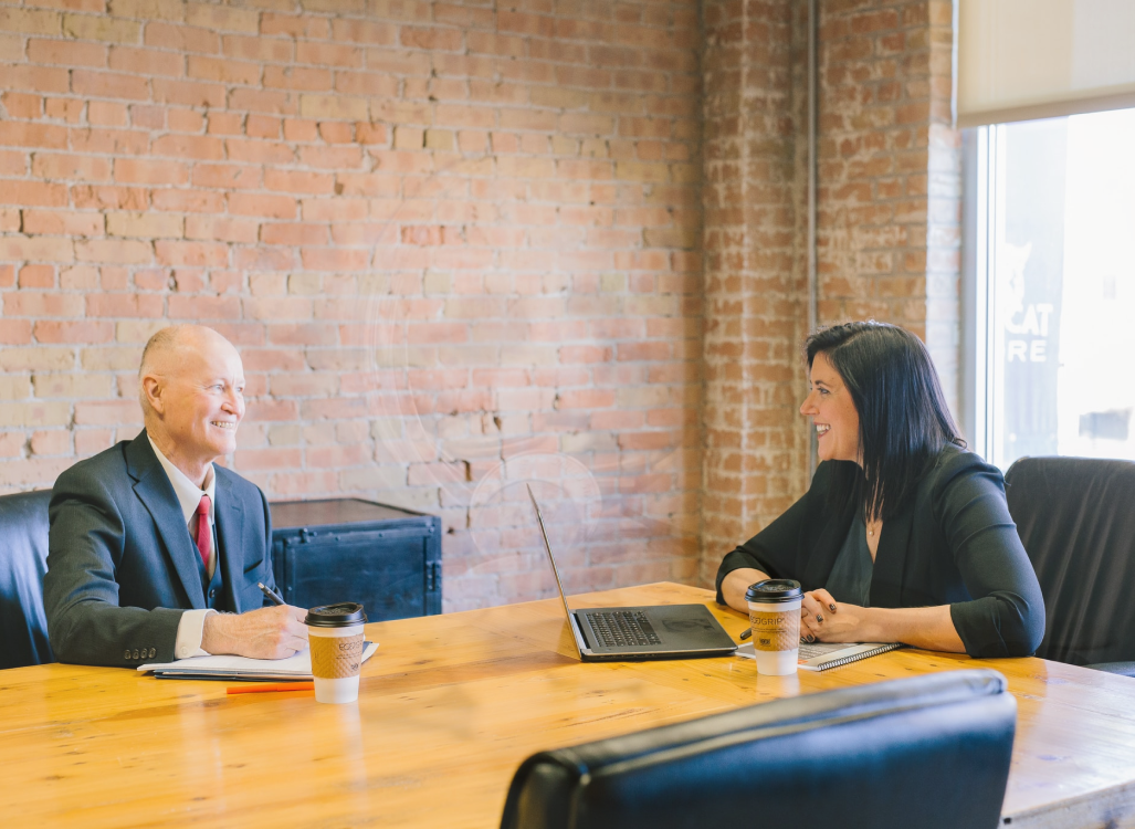 Older man in a suit smiling across a wooden meeting table at a woman with a laptop and coffee cups