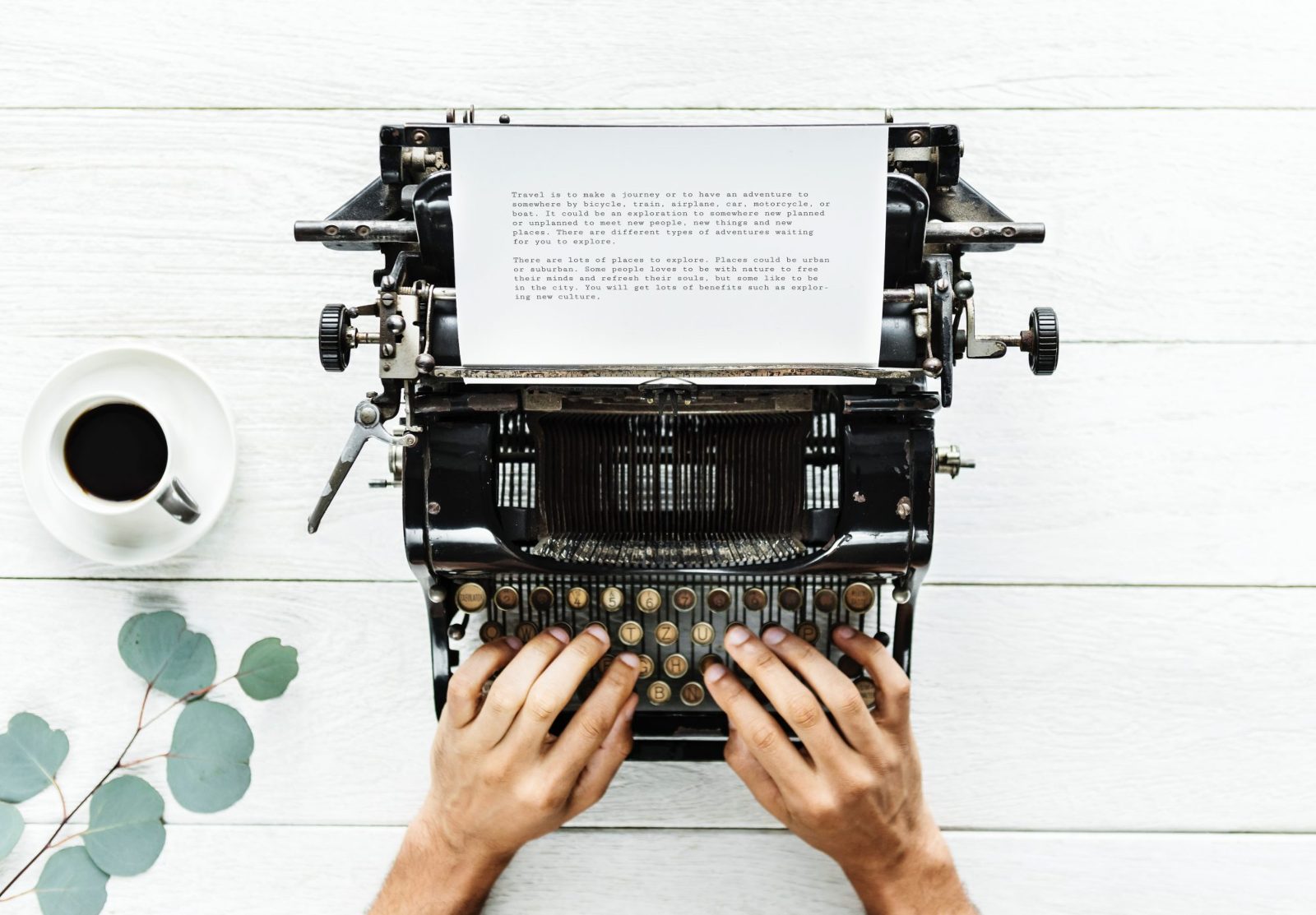 Overhead view of hands typing on a vintage typewriter beside a cup of black coffee on a white desk
