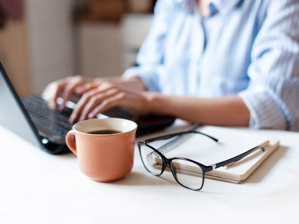 Hands typing on a laptop beside a terracotta coffee cup, glasses and a notebook on a white desk