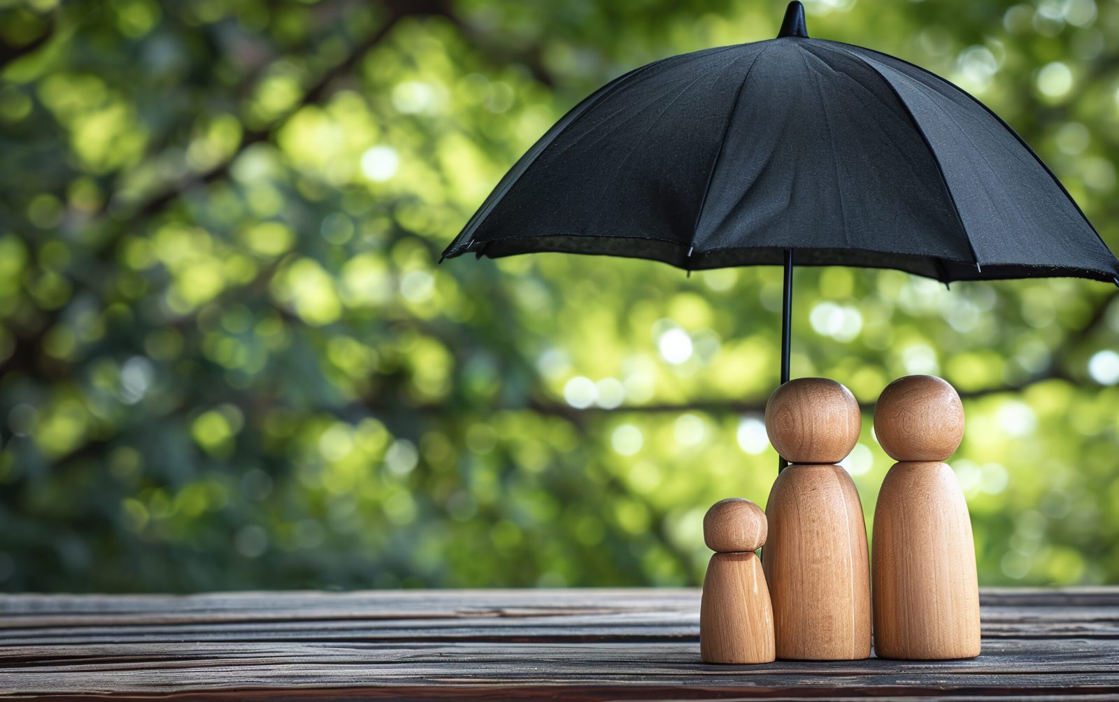 Black umbrella sheltering three wooden family figures on a table against green leafy bokeh