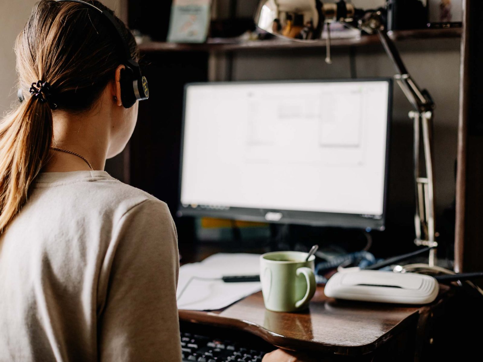 Woman in a headset working at a desktop computer at home, seen from behind, with a mug on the desk