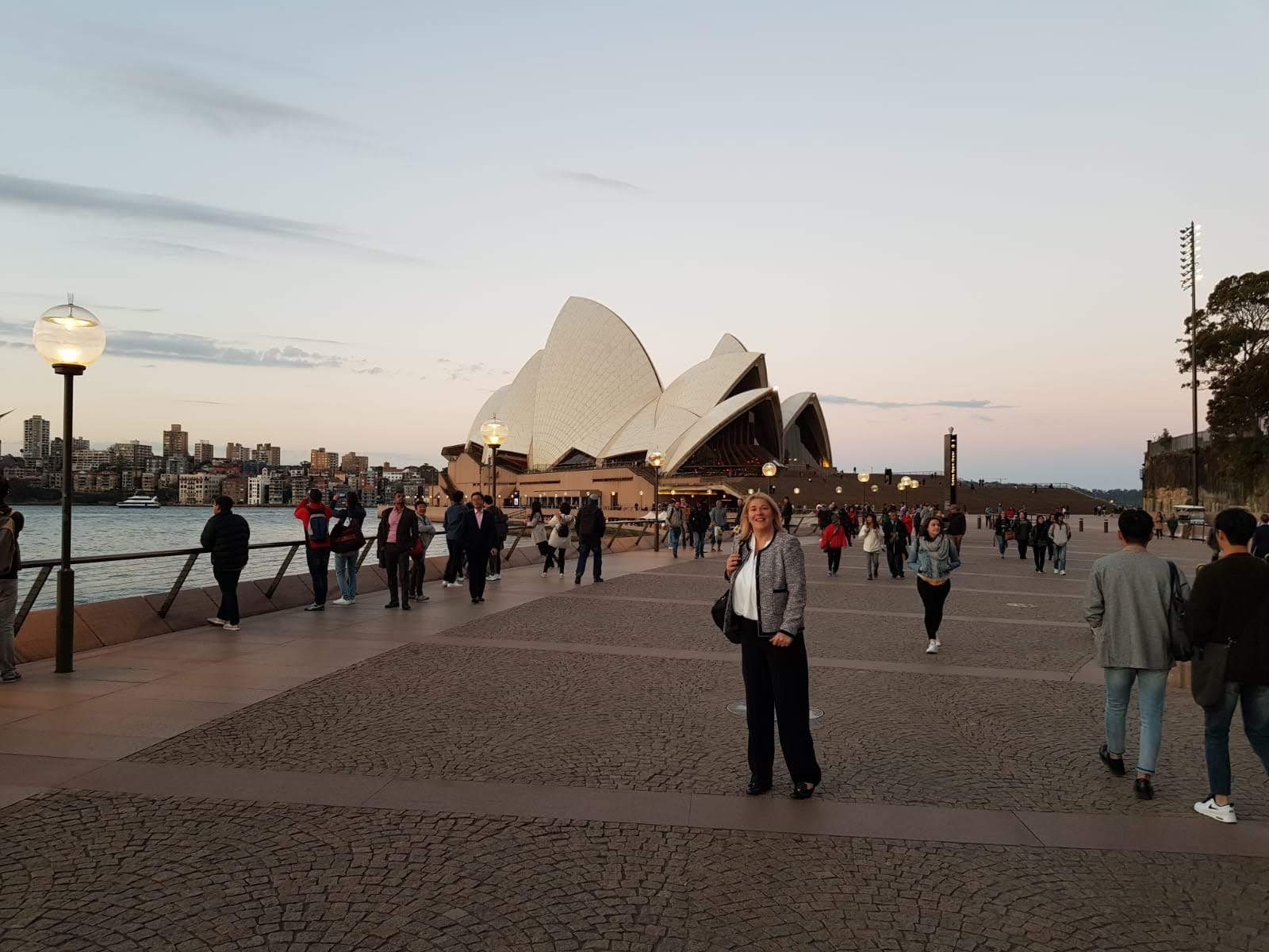 Woman standing on a harbourside promenade with the Sydney Opera House behind at dusk