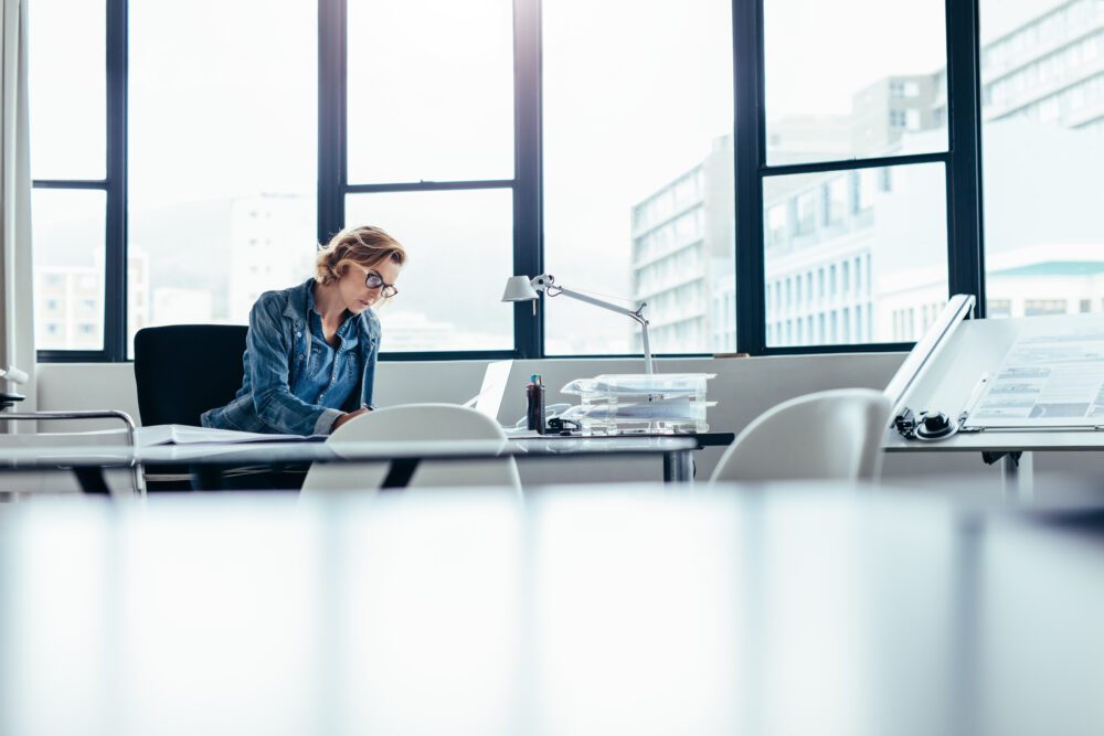 Woman in glasses working alone at a desk in a bright, airy office with tall windows