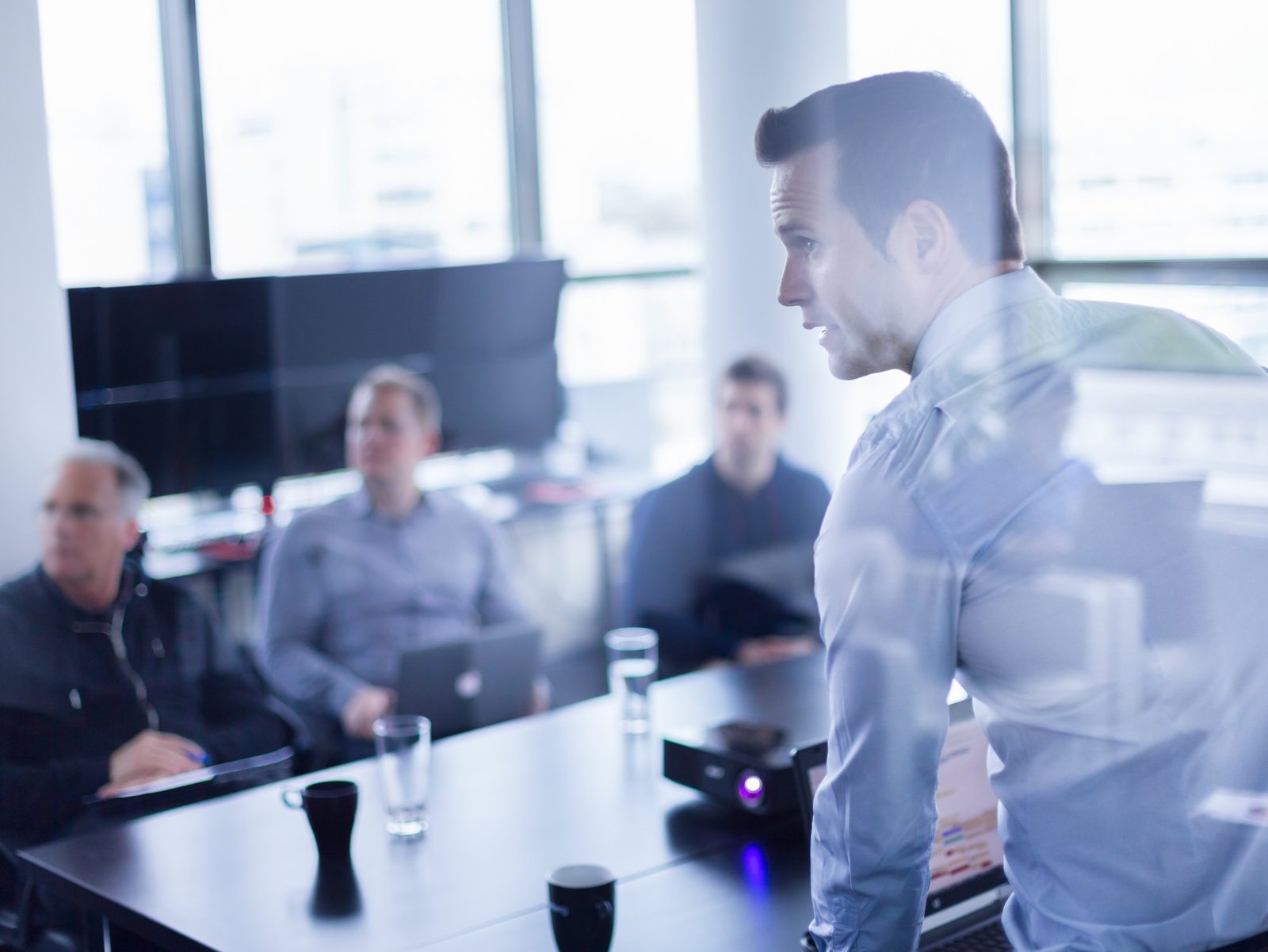 Man standing by a window addressing colleagues seated around a meeting table with laptops