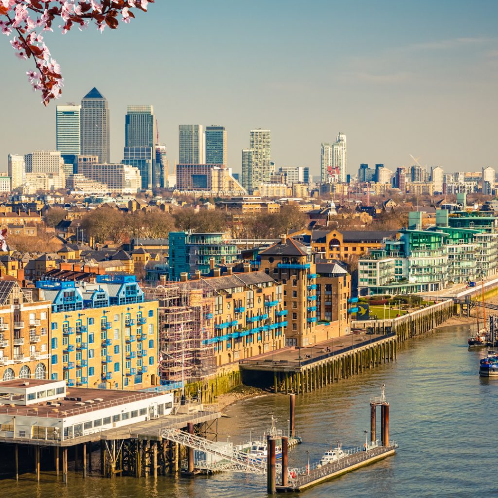 Riverside buildings along the Thames with Canary Wharf towers in the distance and pink blossom in the corner