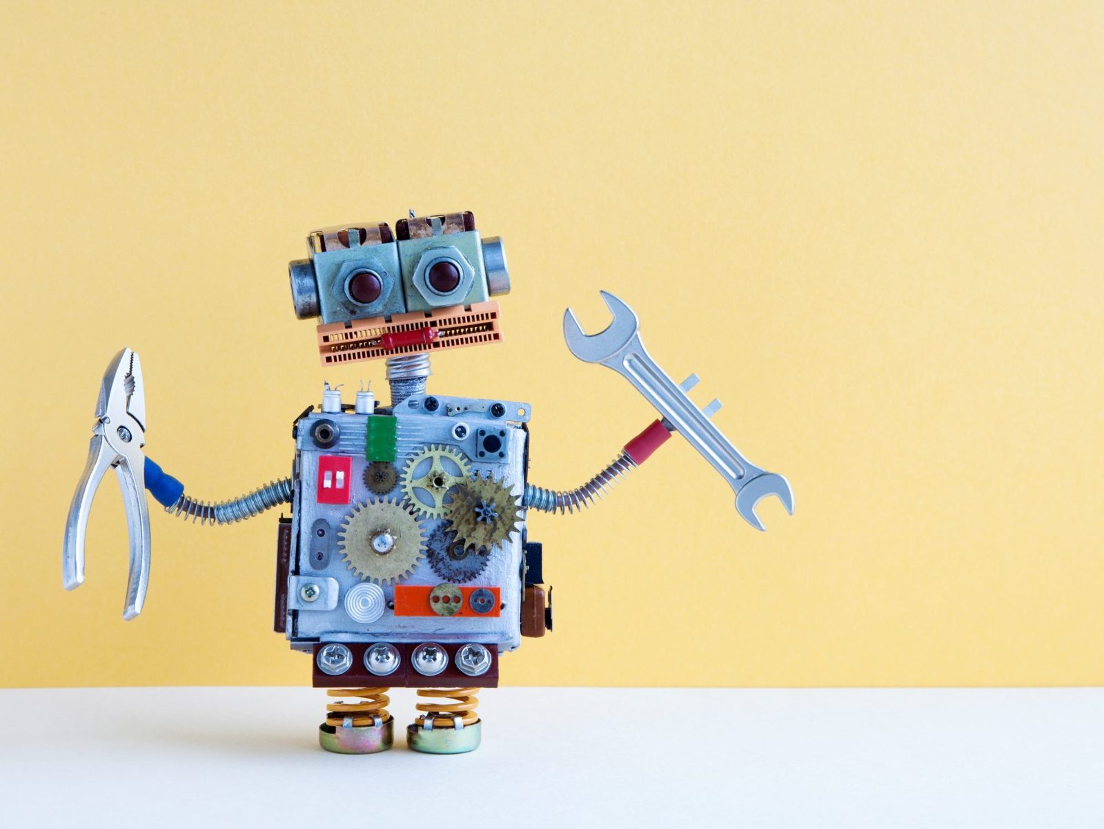 Toy robot built from electronic parts holding pliers and a spanner against a yellow wall