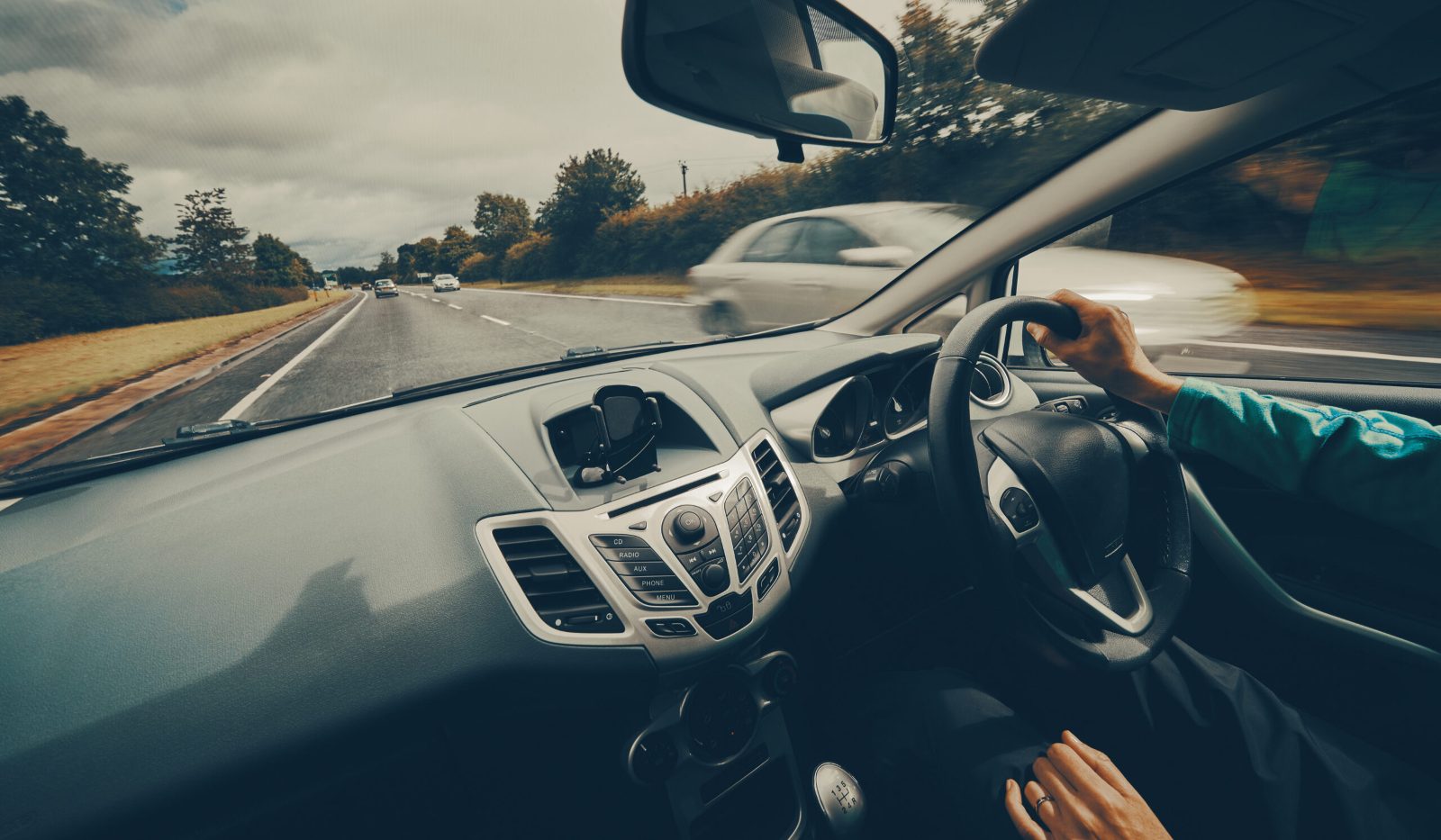 Driver's hands on the steering wheel of a car travelling along a tree-lined road, seen from the passenger seat