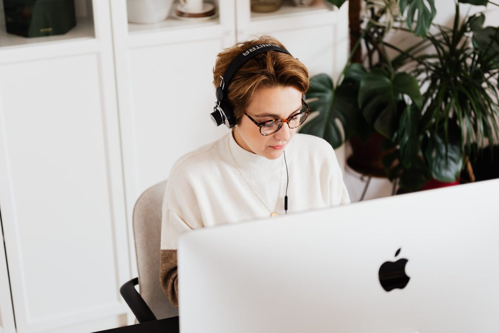 Woman wearing headphones and glasses working at an iMac in a bright home office with plants