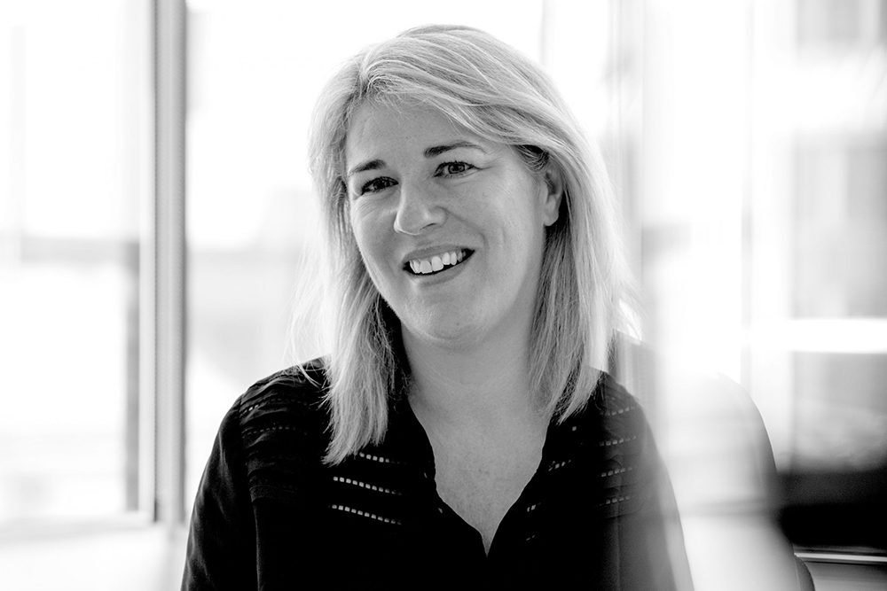 Landscape black-and-white portrait of Joanne Smith smiling in a dark top against a bright window