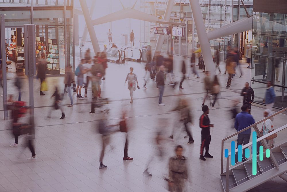 Motion-blurred shoppers crossing a bright, glass-roofed shopping centre concourse, with a blue and teal waveform