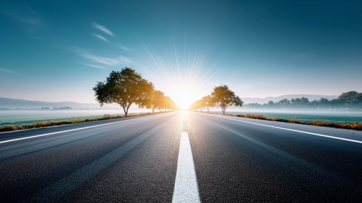 Empty road with a white centre line stretching towards a bright sunrise between two trees