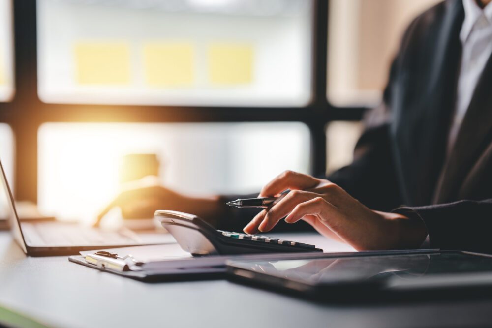 Hands using a calculator and pen beside paperwork and a laptop on a sunlit office desk