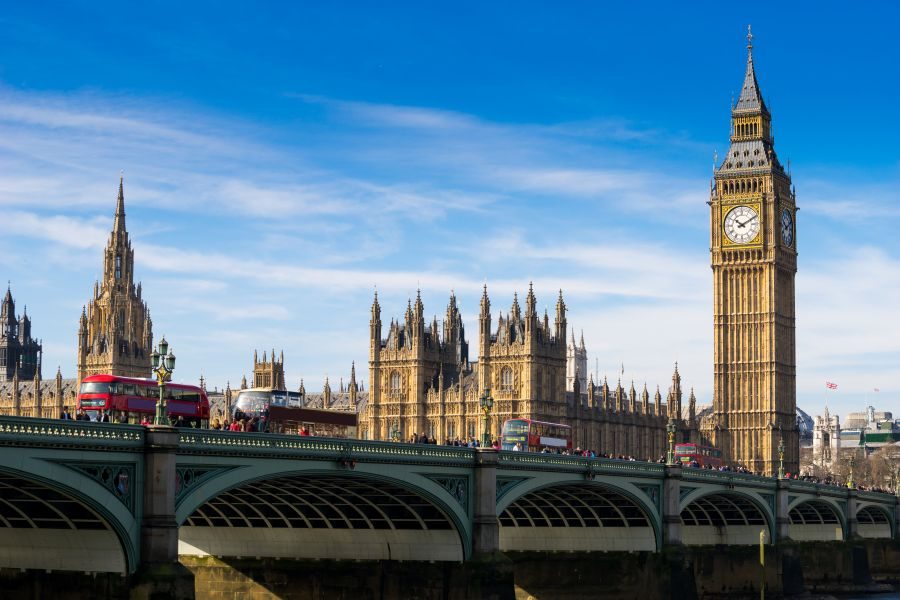 The Houses of Parliament and Big Ben beside Westminster Bridge in London, with red buses crossing under a clear blue sky.
