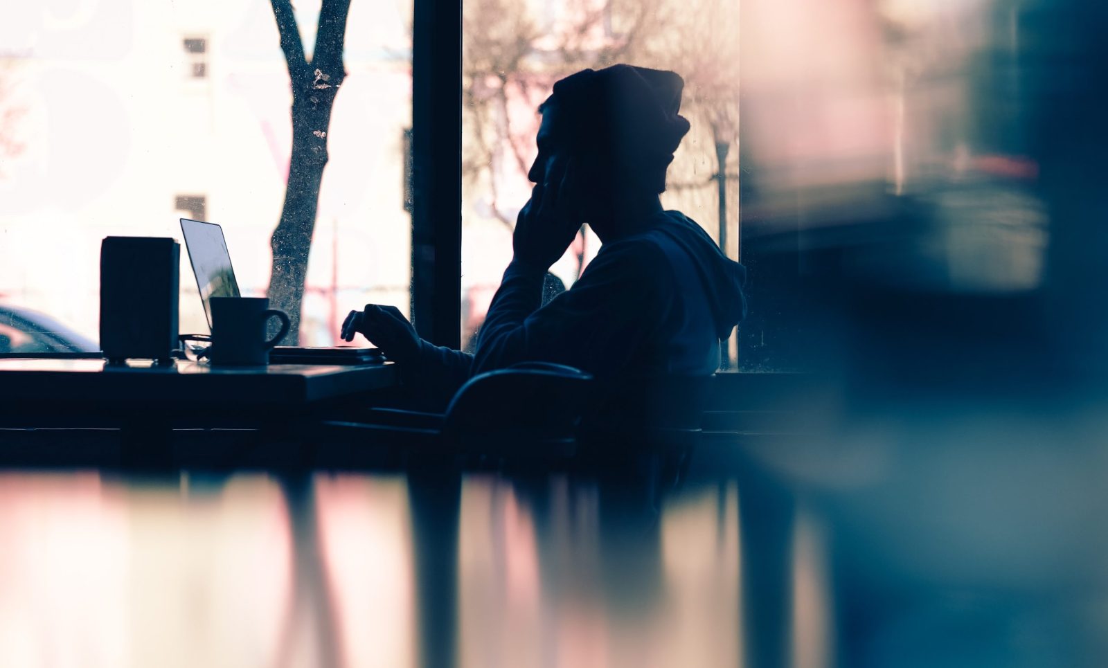 Silhouette of a person in a hood, head in hand, sitting at a laptop by a café window