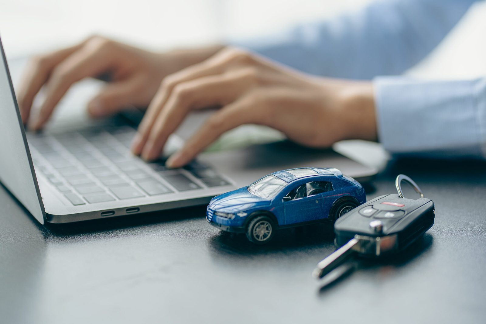 Hands typing on a laptop beside a small blue model car and a car key on a desk