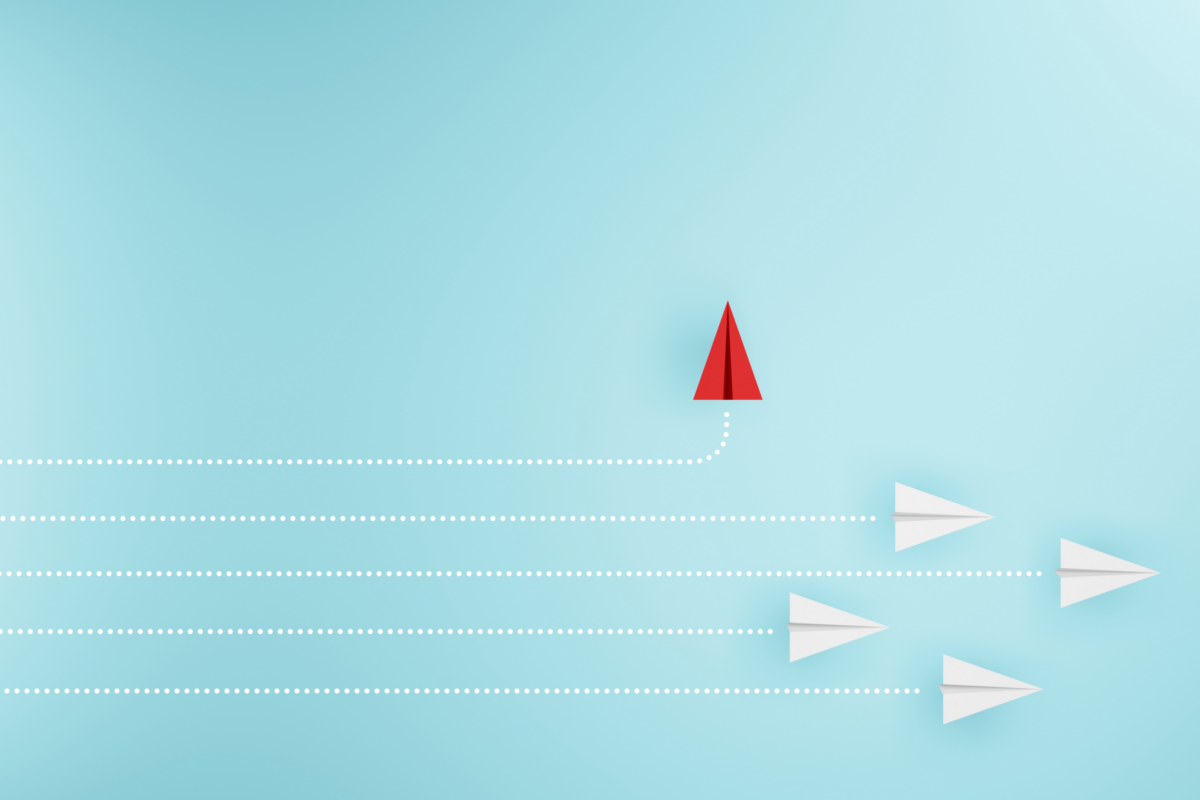 Red paper plane breaking away upwards from a line of white paper planes on a pale blue background