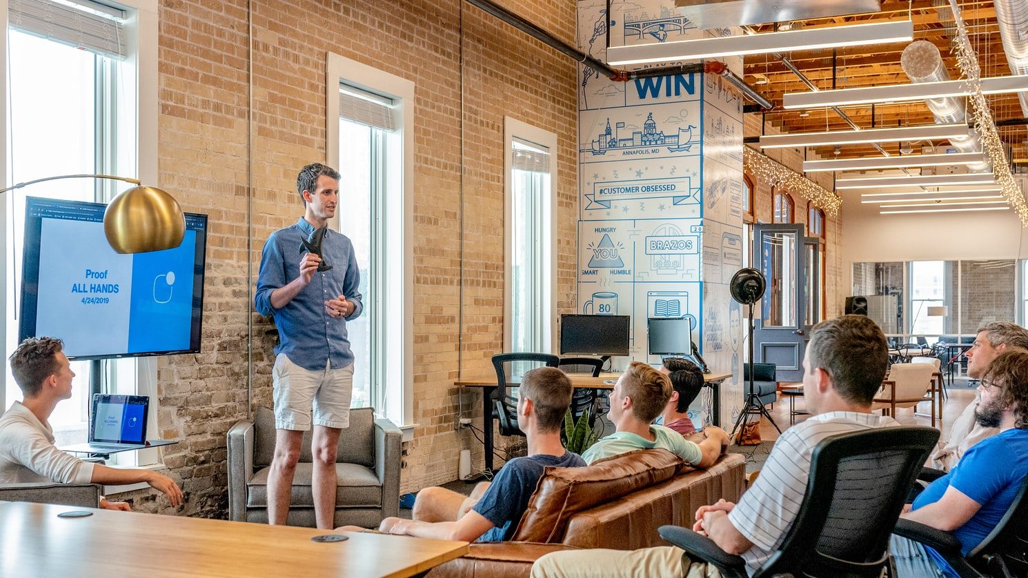 Man presenting to colleagues seated on sofas and chairs in a brick-walled open-plan office
