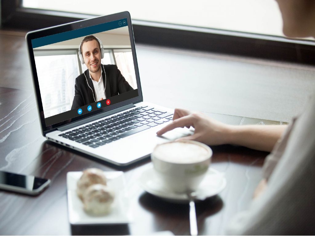 Laptop showing a video call with a smiling man in a headset, beside a cup of coffee on a table