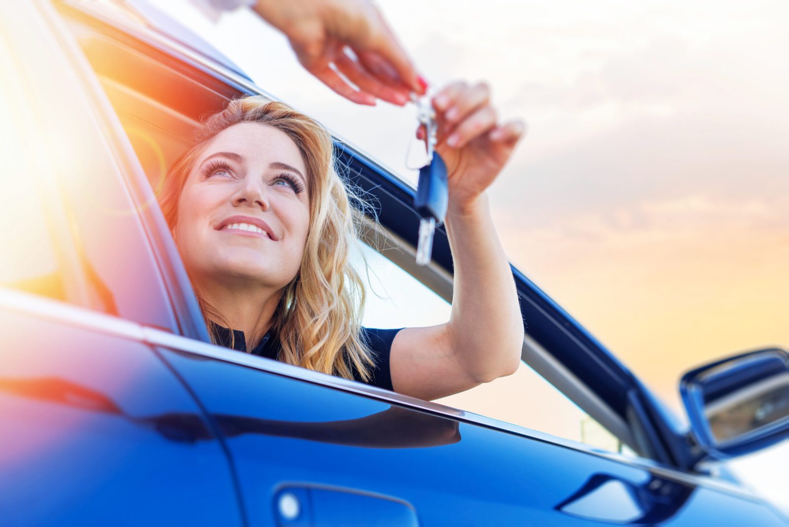 Smiling woman leaning out of a blue car window and holding up a set of car keys against a sunny sky
