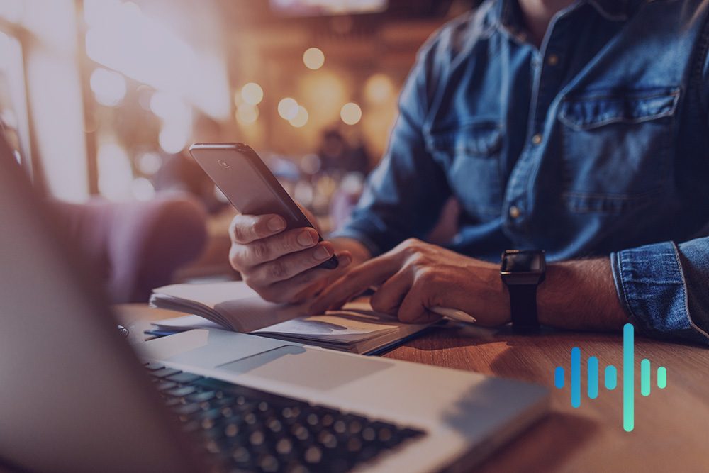 Man in a denim shirt using a smartphone over a laptop and notebook in warm café light, with a blue and teal waveform