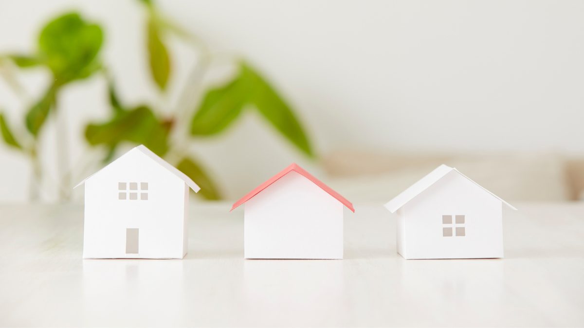 Three small white paper house models, one with a pink roof, on a white table beside a green plant